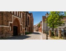 Gepflasterte Gasse in einer Altstadt mit historischen Backsteingebäuden, darunter eine Kirche mit gotischem Spitzbogenportal auf der linken Seite. Rechts sind moderne Wohnhäuser, ein schmiedeeiserner Zaun, ein Baum und eine Straßenlaterne zu sehen.
