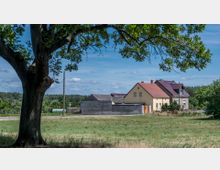 Ein kleines Wohnhaus mit rotem Satteldach und gelber Fassade steht neben einem grauen Haus in einer ländlichen Umgebung. Im Vordergrund ist eine Wiese mit einem großen Baum, im Hintergrund sind Bäume und ein bewaldeter Horizont sichtbar.