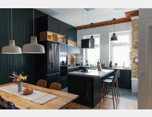 Moderne Küche mit dunklen Einbauschränken und einer zentralen Kochinsel mit Barhockern. Große Fenster lassen Tageslicht herein, eine Wand aus Ziegelsteinen und Holzelemente ergänzen die Einrichtung, während ein Holztisch im Vordergrund steht, dekoriert mit einem Blumenstrauß und einer Obstschale.