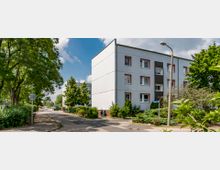 Wohngebiet mit einem dreistöckigen, schlichten weißen Wohnhaus, umgeben von Bäumen, Sträuchern und grüner Vegetation. Im Vordergrund führt ein Bürgersteig entlang der Straße, die von einer Straßenlaterne und einem Verkehrsschild gesäumt ist.