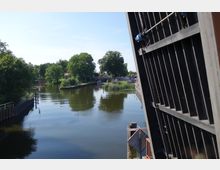 Geöffnete Klappbrücke über einem ruhigen Kanal mit dicht bewachsenen Ufern, Bäumen und einem kleinen Gebäude im Hintergrund. Auf dem Kanal sind Boote und Anzeichen einer Anlegestelle zu sehen.