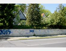Straßenansicht mit einer Mauer, die mit Graffiti bedeckt ist, dahinter ein Garten mit Bäumen und Büschen sowie einem grauen Einfamilienhaus mit Satteldach. Im Vordergrund ist eine asphaltierte Straße mit einem Gehweg sichtbar.