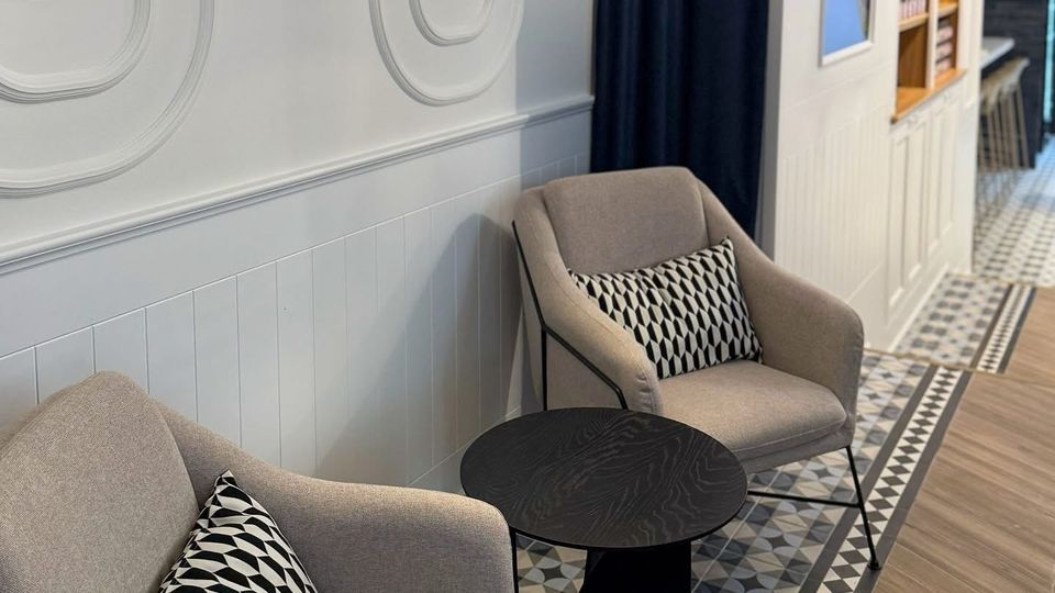 Zwei beige Polstersessel mit gemusterten Kissen stehen neben einem kleinen schwarzen Tisch auf einem gemusterten Fliesenboden. Im Hintergrund befinden sich eine weiße Wand mit integrierten Spiegeln, dunkle Vorhänge und ein Regal mit dekorativen Elementen.