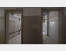 Verlassene Innenansicht eines Gebäudes mit zwei offenen Türrahmen. Die Räume zeigen abblätternde Wände, teilweise weiße Fliesen und sichtbare Leitungen an der Decke.