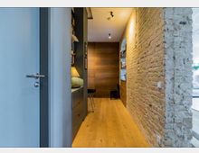 Flur in einem modernen Innenraum mit Holzboden, einer unverputzten Backsteinwand auf der rechten Seite und eingebauten Regalen mit Büchern. Links befindet sich eine Schrankwand mit einer Tischlampe, und im Hintergrund ist eine Holzverkleidung sichtbar.