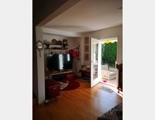 Wohnzimmer mit hellen Holzfußböden, einem großen Fernseher auf einem Sideboard und verschiedenen Dekorationsgegenständen an der Wand. Rechts führt eine geöffnete Glastür auf eine Terrasse mit Blick auf Gartenmöbel und grüne Hecken im Außenbereich.