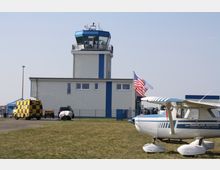 Flugplatz mit Kontrollturm, einem kleinen weißen Flugzeug im Vordergrund und einer amerikanischen Flagge neben dem Gebäude. Im Hintergrund stehen ein gelb-schwarz karierter Transporter und weitere Fahrzeuge.