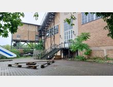Backstein-Gebäude eines ehemaligen Industriekomplexes mit großen Fenstern, einer Metalltreppe und umliegenden Pflanzen. Im Vordergrund gepflasterter Hof mit alten Holzgestellen und Metallkonstruktionen.