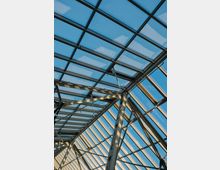 Glasdachkonstruktion mit einem Metallrahmen, durch den blauer Himmel sichtbar ist. Die Architektur zeigt diagonale Stahlstreben und rechtwinklige Glaselemente.