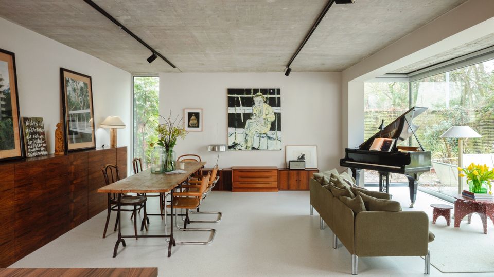 Open-plan modern living and dining room with a raw concrete ceiling, white walls, and track lighting, featuring a long wooden dining table, built-in wood cabinetry, and framed artwork. Large floor-to-ceiling windows overlook a garden, with a grand piano and seating area near the glass.