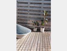 Holz-Terrasse mit einer grauen, freistehenden Metallbadewanne auf der linken Seite, zwei Topfpflanzen auf der rechten Seite und einer Sichtschutzwand aus horizontal gestapelten Holzlatten im Hintergrund.