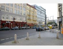 Straßenszene in einer Stadt mit mehrstöckigen Gebäuden im historischen Stil links und moderner Architektur rechts. Geparkte Autos säumen die Straße, und im Vordergrund ist ein Gehweg mit Pollern sichtbar.