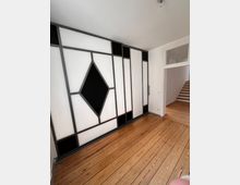 Raum mit Holzdielenboden und einer Wand aus geometrischen Elementen in Schwarz-Weiß, darunter ein auffälliges rautenförmiges Muster. Im Hintergrund ist eine Tür mit Glaseinsatz sowie eine Treppe zu sehen.