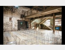 Industrieller Veranstaltungsraum mit unverputzten Backsteinwänden, einer Metalltreppe und einer offenen Galerie. Klarsichtstühle sind in Reihen aufgestellt, und ein Schild mit der Aufschrift „Sofia“ steht auf einem Ständer.