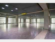 Offener, moderner Innenbereich eines Parkhauses mit Betonwänden und -säulen, glatten Böden und beleuchteter Rasterdecke. Die Wände sind mit matten Glasfenstern versehen, die Licht durchlassen.