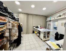 Ein heller Arbeitsraum mit Fliesenboden, Kleiderstange mit Stoffen und Kleidungsstücken links, einem großen Arbeitstisch mit Materialien in der Mitte und Regalen an den Wänden. Im Hintergrund befinden sich Vorhänge, die den Raum abtrennen, sowie ein Fenster mit einer Leiter daneben.