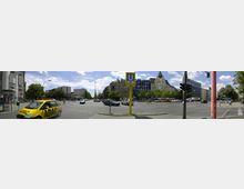 Straßenkreuzung in einer Stadt mit mehreren Fahrzeugen, darunter ein gelbes ADAC-Auto. Im Hintergrund stehen Gebäude mit unterschiedlichen Fassaden, Bäume säumen die Straße, und ein gelbes U-Bahn-Schild kennzeichnet einen nahegelegenen U-Bahn-Eingang.