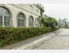 Seitenansicht eines historischen Gebäudes mit Rundbogenfenstern und leicht verwitterter Fassade, bewachsen von Kletterpflanzen. Vor dem Gebäude verläuft eine gepflasterte Straße, daneben eine dichte Hecke und im Hintergrund ist eine kleine Brücke zu sehen.