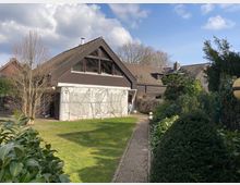 Zweistöckiges Wohnhaus mit dunklem Satteldach und heller Fassade, umgeben von einem gepflegten Garten mit Rasen, Hecken und einem schmalen gepflasterten Weg. Im Hintergrund sind weitere Häuser sowie Bäume unter einem teils bewölkten Himmel sichtbar.