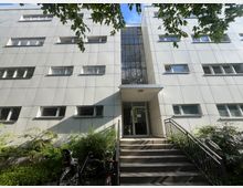 Eingangsbereich eines modernen, mehrstöckigen Gebäudes mit hellgrauer Fassadenverkleidung und Reihen von rechteckigen Fenstern. Eine Treppe mit Geländer führt zu einer verglasten Tür unter einem kleinen Vordach, umgeben von Büschen und überhängenden Baumzweigen.