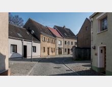 Eine kleine, gepflasterte Straße mit niedrigen Wohnhäusern in einem ländlichen oder dörflichen Umfeld. Die Gebäude haben einfache Fassaden aus Putz in Beige- und Pastelltönen, teils mit Dachfenstern, und werden von vereinzelten Straßenlaternen gesäumt.