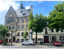 Historisches Gebäude mit verzierten Giebeln und Uhr auf der linken Seite, neben einem niedrigeren, schlichten Wohnhaus. Im Vordergrund eine Straße mit geparkten Autos, Fußgängern und Bäumen.