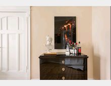 Eine helle Wand mit einer weißen Tür links, daneben eine glänzende schwarze Kommode mit goldenen Griffen, auf der sich eine Büste, einige Flaschen alkoholischer Getränke und ein gerahmtes Bild mit einer nächtlichen Szene befinden.