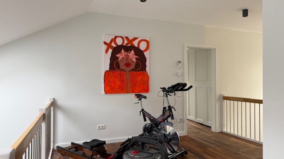 Upstairs loft landing with sloped white ceilings, dark wood flooring, and a wooden railing along the edge. A stationary exercise bike and rowing machine sit beneath a large wall painting, with a white door and hallway opening to the right.