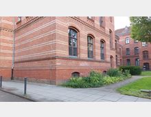 Ziegelgebäude mit mehrstöckiger Fassade, gekennzeichnet durch dekorative Muster aus roten und hellen Ziegeln sowie große, bogenförmige Fenster. Im Vordergrund führt ein gepflasterter Gehweg an einem Beet mit bunten Pflanzen entlang, während im Hintergrund ein grüner Rasen und weitere Gebäudeteile sichtbar sind.