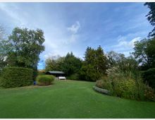 Ein weitläufiger Garten mit einer gepflegten Rasenfläche, umgeben von hohen Bäumen, dichten Sträuchern und Zierpflanzen. Im Hintergrund steht eine kleine Holzhütte mit einer weißen Bank davor, unter einem bewölkten blauen Himmel.