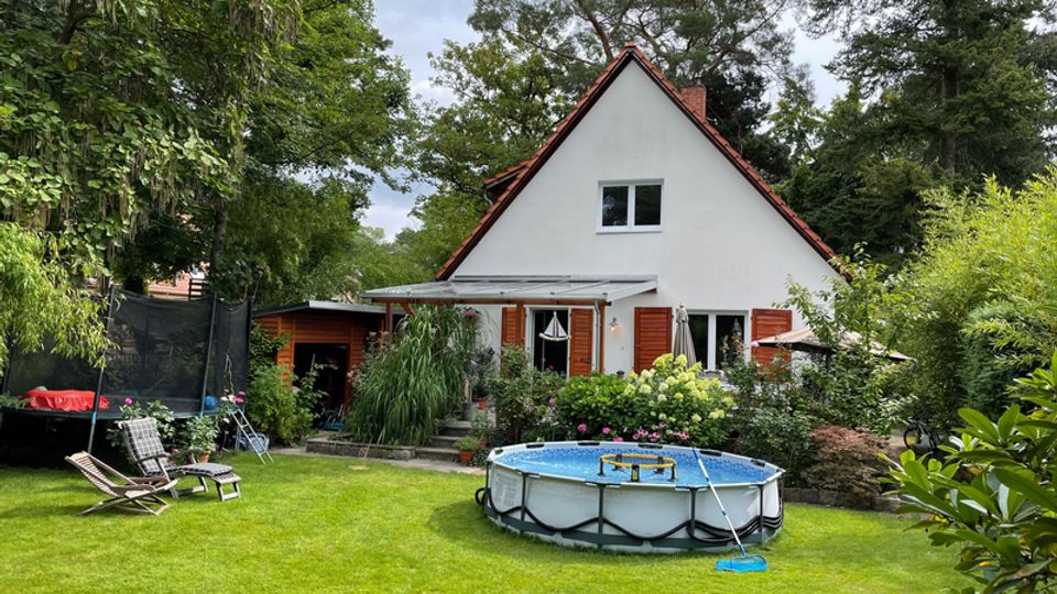 Einfamilienhaus mit steilem Satteldach und weißen Fassaden, umgeben von dichtem Baumbestand und Gartensträuchern. Im Vordergrund liegt eine gepflegte Rasenfläche mit rundem Aufstellpool, Liegestühlen und einem Trampolin am Rand.
