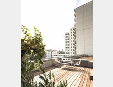 Holzterrasse auf einem Dach mit modernem Sitzbereich aus weißen Metallmöbeln, umgeben von Pflanzen. Im Hintergrund sind moderne Wohn- und Bürogebäude zu sehen.