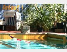 Außenbereich eines Schwimmbades mit einem Pool und umgebender Vegetation; im Hintergrund stehen ein Strandkorb, Pflanzen und eine Glaswand mit der Aufschrift „Anlauf“.