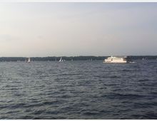 Ein großer See mit einer weißen Passagierfähre im Vordergrund und mehreren kleinen Segelbooten im Hintergrund. Am Horizont ist eine Baumreihe zu sehen, unter einem leicht bewölkten Himmel.