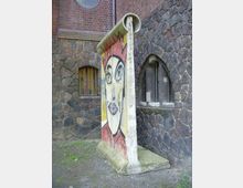 Ein aufrecht stehendes Segment der Berliner Mauer mit bunter Graffiti-Kunst, das ein Gesicht darstellt, vor einer Fassade aus Ziegel- und Naturstein mit kleinen, bogenförmigen Fenstern. Der Boden ist grasbewachsen und wirkt leicht verwittert.