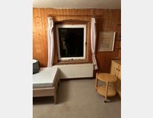 Holzgetäfeltes Zimmer mit einem Fenster in der Mitte, weißen Vorhängen, einem Heizkörper darunter sowie einem Bett links und einem kleinen runden Beistelltisch rechts. Rechts an der Wand hängen ein gerahmtes Bild und ein Regal. Der Boden ist mit einem Teppich ausgelegt.