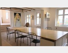 Heller Raum mit modernem Esstisch aus Holz und schwarzen Stühlen, mehreren Fenstern mit Blick nach draußen und Holzbalken an der Decke. An der Wand hängt ein großes Porträtgemälde, daneben stehen ein antiker Sessel und minimalistische Lampen.