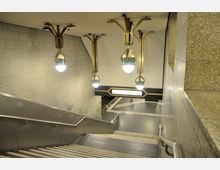 Ansicht eines U-Bahn-Eingangs mit einer Treppe, die zu einer unterirdischen Plattform führt. Die Umgebung ist mit hellen Steinwänden gestaltet, und mehrere auffällige goldene Leuchten mit runden Glühbirnen hängen an der Decke; im Hintergrund ist ein beleuchtetes Stationsschild erkennbar.