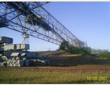 Großer Schaufelradbagger in einer Tagebau-Umgebung, mit massiven Metallträgern, Fördermechanismen und Gleisen im Vordergrund; umgeben von grasbedeckten Hügeln unter klarem Himmel.