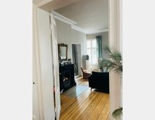 Wohnzimmer in einer Altbauwohnung mit hellen Wänden, Holzdielenboden und Stuckverzierungen an der Decke. Im Raum befinden sich ein schwarzer Kamin mit Dekoration, ein Sofa, Sessel, ein großer Spiegel und ein Fenster mit Vorhängen, das natürliches Licht einlässt.