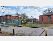 Industrielle Umgebung mit Backsteingebäuden, einem grünen Tor mit der Beschriftung "Tor 9" und einer gelben Lokomotive dahinter. Vor dem Gelände befindet sich ein Bahnübergang mit Schranken und einem Andreaskreuz.