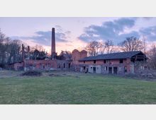 Ruinen eines alten Backsteingebäudes mit verfallenen Dächern und einem hohen Schornstein, umgeben von einem offenen Gelände mit Rasen und Bäumen im Hintergrund bei Sonnenuntergang.