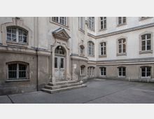 Innenhof eines historischen Gebäudes mit beigefarbener Fassade, verzierten Fensterrahmen und einem zentralen Eingang mit Doppeltüren, geschmückt durch ein kleines Giebeldach. Der Boden besteht aus grauem Asphalt, und die Mauern zeigen Spuren von Alterung.