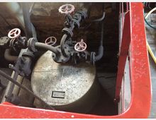 Industrieanlage mit metallischem Kessel, mehreren verbundenen Rohren und Ventilen mit roten Handrädern. Der Raum zeigt eine Backsteinwand im Hintergrund und hat teilweise sichtbare, abgenutzte Oberflächen.