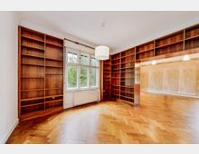 Zimmer mit Holzparkettboden und eingebauten, raumhohen Bücherregalen aus dunklem Holz. An der Wand befindet sich ein großes Fenster mit Blick auf grüne Bäume, darunter ein Heizkörper; daneben ein Durchgang zu einem weiteren Raum.