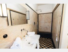 Modernes Badezimmer mit beigen Natursteinfliesen und schwarzer Rahmenkante, großem Wandspiegel und Waschbecken im Vordergrund. Hinten befindet sich eine Dusche mit Wand- und Handbrause sowie ein schwarz-weiß kariert gefliester Boden.