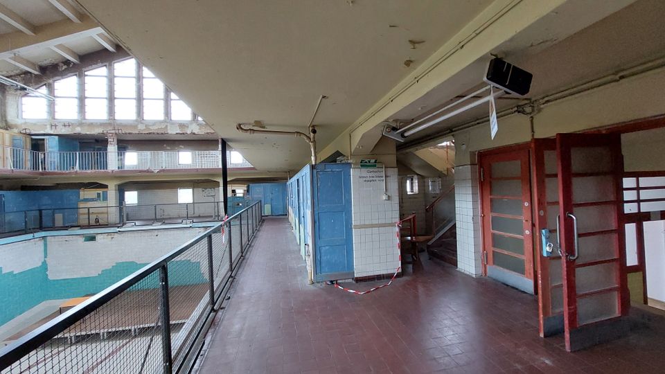 Innenansicht eines alten, verlassenen Schwimmbads mit einer leeren Halle und abgenutzten Umkleidekabinen. Die Umgebung zeigt Fliesenwände, Metallgeländer und Fenster mit viel Tageslicht sowie beschädigte Oberflächen.