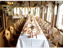 Rustikal eingerichteter Gastraum mit einer langen, festlich gedeckten Tafel, umgeben von hölzernen Stühlen mit geschwungenen Lehnen. Große Fenster mit weißen Vorhängen lassen Tageslicht in den Raum, der mit rustikalen Dekorationselementen wie Jagdtrophäen und antiken Lampen geschmückt ist.