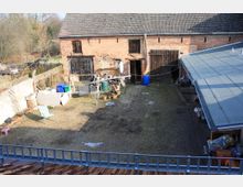 Ein ländlicher Hof mit einem gepflasterten und grasbewachsenen Innenhof. Auf der linken Seite steht ein aus rotem Backstein gebautes, altes Gebäude mit Holztüren, rechts ein Gebäude mit einem grauen Flachdach. Im Hof gibt es diverse Gegenstände wie Gartenmöbel, Pflanzkübel und Abfallbehälter.