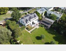 Zweistöckiges, weißes Wohnhaus mit dunklem Satteldach und großem Garten mit gepflegtem Rasen, umgeben von Bäumen und Hecken. Im Hintergrund sind weitere Gebäude sowie eine Zufahrtsstraße mit parkenden Autos sichtbar.